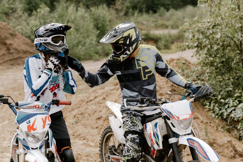 Two motocross riders on dirt bikes gearing up on a trail outdoors.